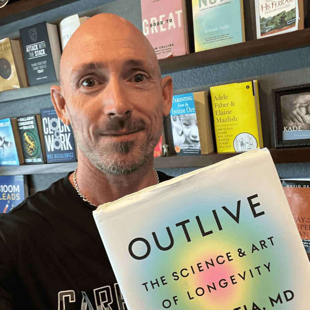 OrthoHeaven A person holding the book "Outlive: The Science & Art of Longevity" in front of a bookshelf filled with various other books, creating an inspiring OrthoHeaven for avid readers. Freedom Mapping