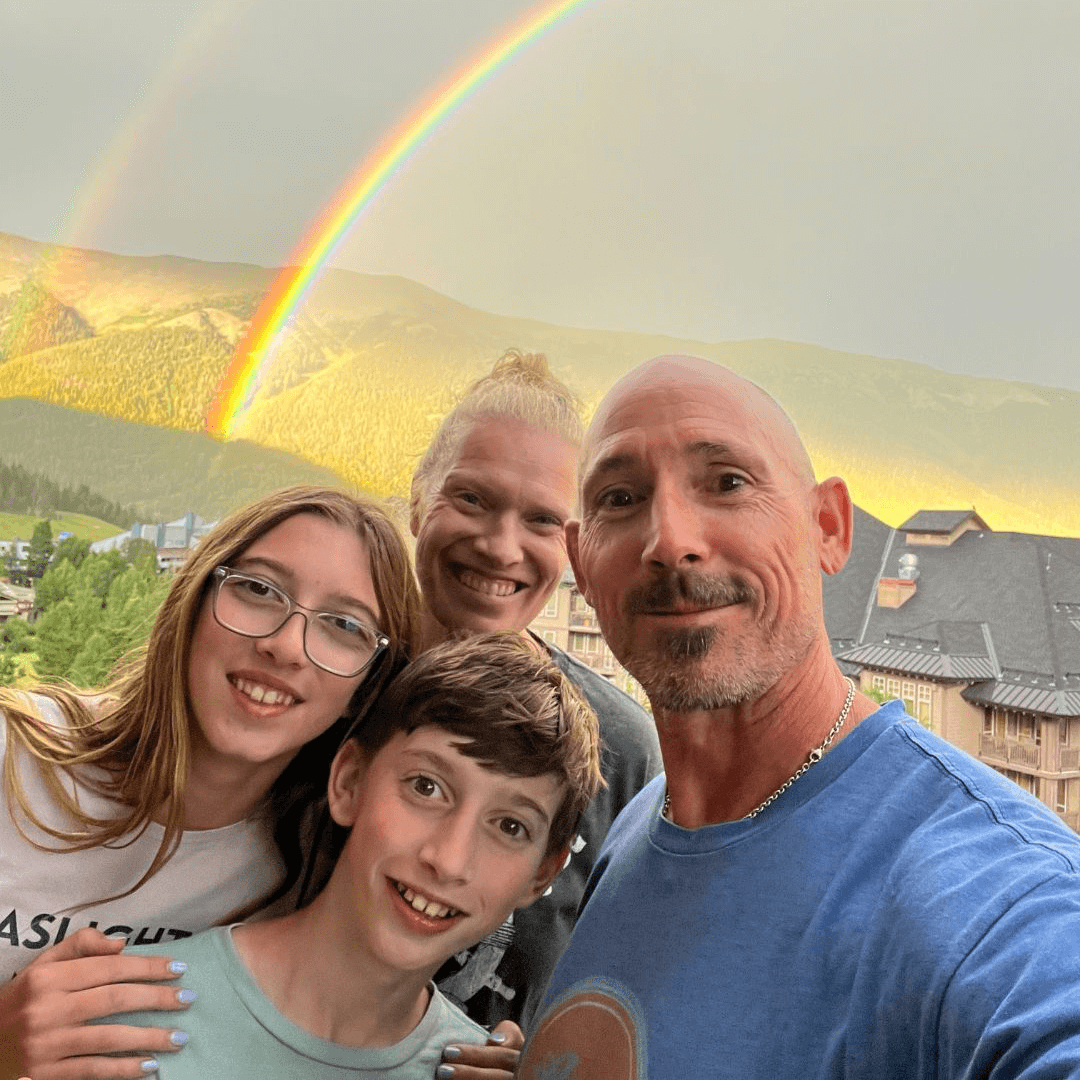 OrthoHeaven Four people pose for a selfie outdoors with mountains, trees, and a bright double rainbow in the background, capturing an OrthoHeaven moment of pure joy and natural beauty. Freedom Mapping