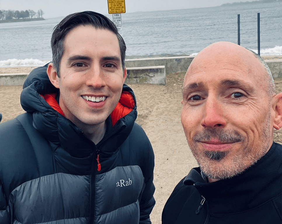 OrthoHeaven Two men stand close together and smile at the camera with OrthoHeaven hats on, a lake, sandy shore, and overcast sky in the background. Freedom Mapping