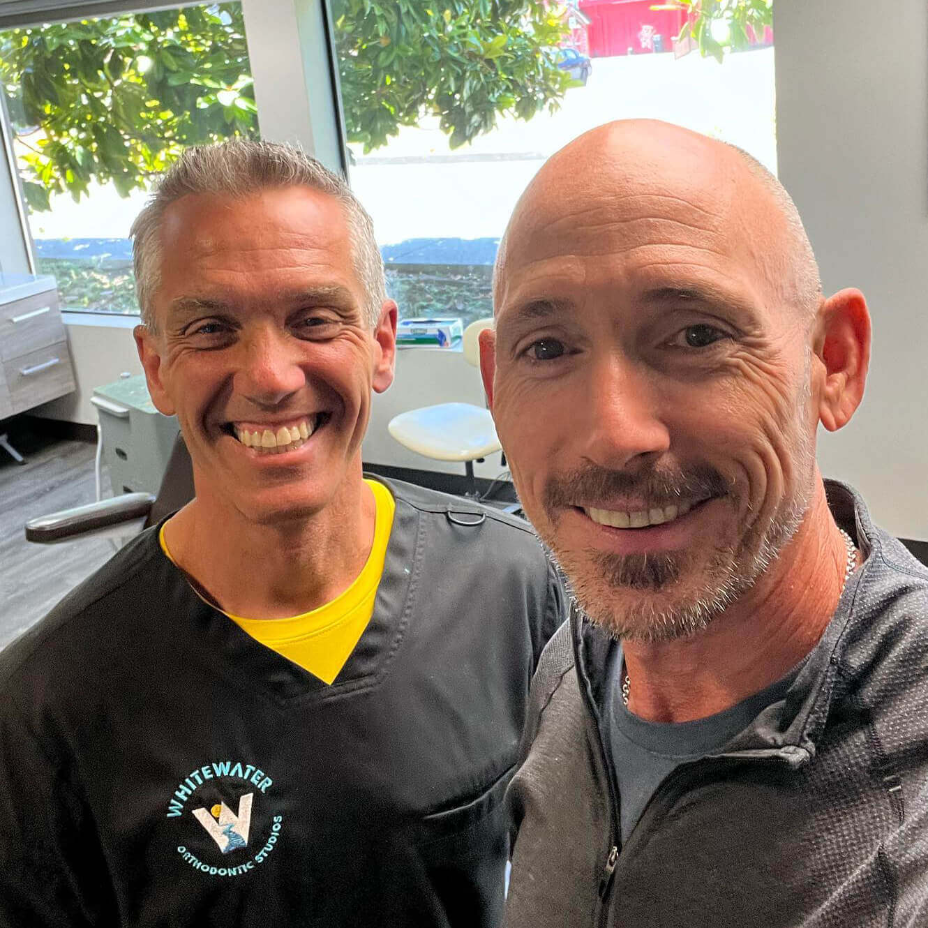 OrthoHeaven Two men smiling and posing for a selfie in a bright office, one wearing dental scrubs with the Whitewater Orthodontic Studios logo, capture an OrthoHeaven moment. Freedom Mapping