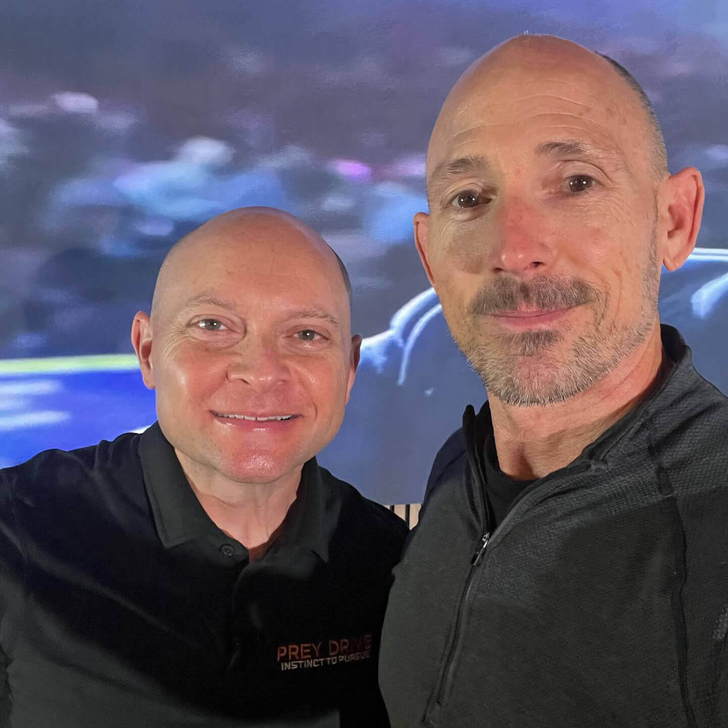 OrthoHeaven Two men with shaved heads stand close together indoors, facing the camera and smiling. One man wears an OrthoHeaven black polo shirt, the other a black zip-up jacket. A blurred screen is visible in the background. Freedom Mapping