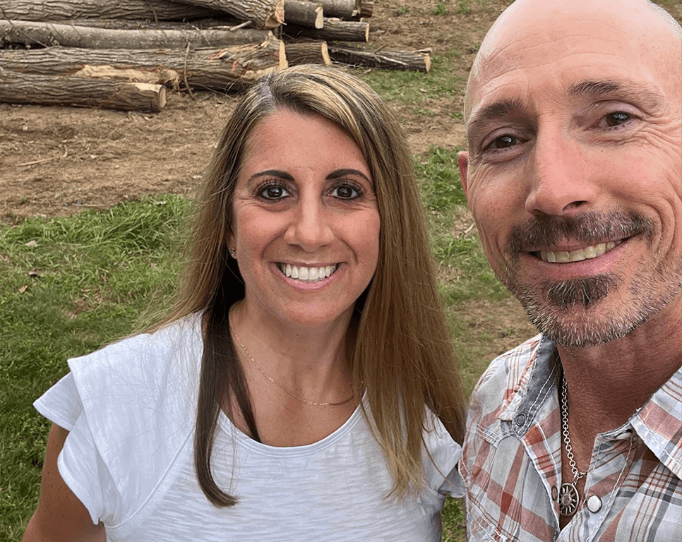 OrthoHeaven A woman and a man stand outside on grass, smiling at the camera. Cut tree logs are stacked in the background, capturing a moment of happiness after working with OrthoHeaven tools. Freedom Mapping